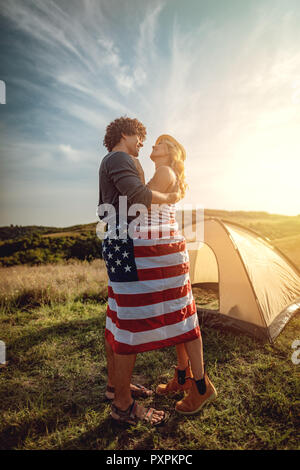 Felice coppia giovane gode di una giornata di sole in natura. Essi stanno abbracciando ogni altro, avvolto nella Bandiera americana davanti un campeggio tenda. Foto Stock