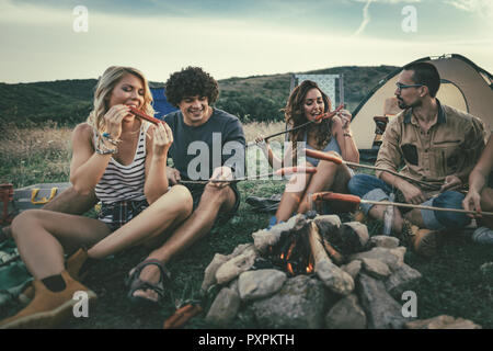 Felice giovani amici di godere di una giornata di sole in montagna. Stanno ridendo e tostatura di salsicce su bastoni su un accampamento vicino alla tenda. Foto Stock