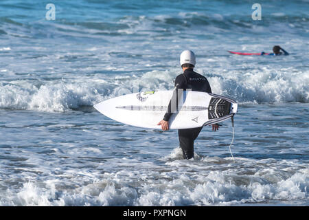 Un surfista che indossa un casco di sicurezza e portando la sua tavola da surf a piedi in mare a Fistral Beach in Newquay in Cornovaglia. Foto Stock