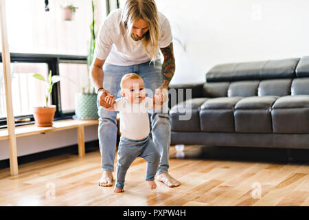 Padre aiutare la nostra bambina prendere i primi passi a casa Foto Stock