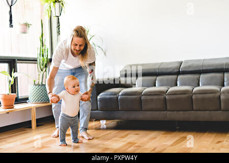 Padre aiutare la nostra bambina prendere i primi passi a casa Foto Stock