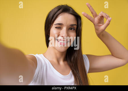 Piacevole attraente ragazza rendendo selfie in studio e ridere. Bella giovane donna con capelli castani sta immagine di se stessa il colore giallo brillante background. Foto Stock