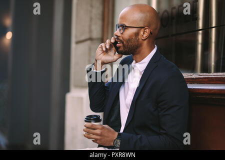Felice uomo d'affari africano seduto fuori a parlare al telefono con una tazza di caffè. uomo d'affari che si rilassa all'aperto facendo una telefonata. Foto Stock