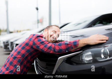 Un Felice giovane uomo abbraccia il suo auto al di fuori del garage negozio Foto Stock