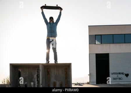 Guidatore di skateboard vestito in abiti casual sulla parte superiore di una scatola di legno con lo skateboard sopra la sua testa Foto Stock