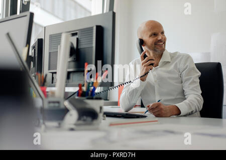 Imprenditore sorridente seduto alla sua scrivania che parla al telefono Foto Stock