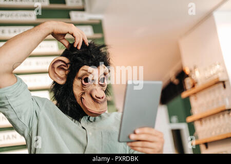 Uomo che indossa la maschera di scimmia utilizzando digitale compressa, graffiare la testa Foto Stock