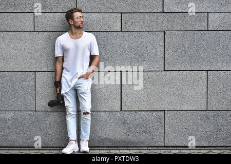 Foto del bel ragazzo caucasico su grigio Parete strutturata in T-shirt bianco e bianco jeans con fotocamera appesa sulla fascetta sul suo braccio. Foto Stock