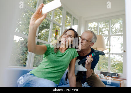 Felice Coppia matura prendendo un selfie a casa con uomo giocattolo di suonare una chitarra elettrica Foto Stock