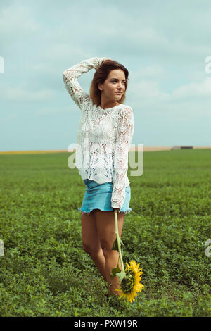 Giovane donna tenendo un girasole, stando in piedi in un campo verde Foto Stock