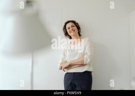 Imprenditrice appoggiata contro la parete in ufficio con le braccia incrociate, sorridente Foto Stock