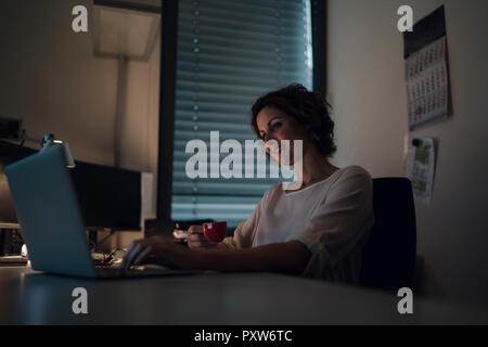 Imprenditrice lavoro straordinario, utilizzando laptop, bere caffè Foto Stock