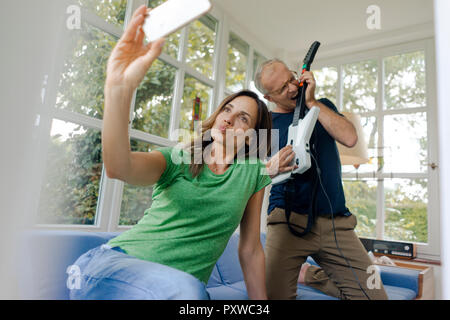 Donna matura prendendo un selfie a casa con uomo giocattolo di suonare una chitarra elettrica Foto Stock