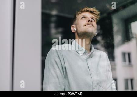 Giovane uomo in piedi alla finestra tramite auricolari, fantasticando Foto Stock