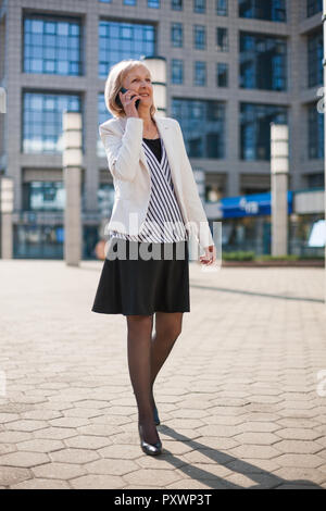 Felice senior imprenditrice sta parlando al telefono al di fuori dell'edificio dell'azienda. Foto Stock