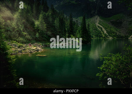 Vista panoramica su gli escursionisti sul sentiero in alta montagna con il lago accanto a loro e raggi di sole che cadono dagli alberi. Foto Stock