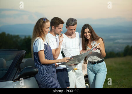 Quattro amici, due ragazze e due ragazzi pianificazione viaggio in automobile. Azienda in piedi vicino a cabriolet, sorridente, azienda mappa e lo sta guardando. Giovani viaggiatori a discutere del percorso di viaggio. Foto Stock