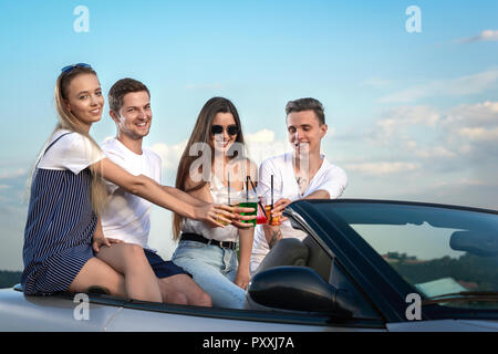 Società di quattro amici seduti insieme su cabriolet e bicchieri tintinnanti con limonata. Belle ragazze e giovani uomini belli divertendosi e godendo di avventure estive. Foto Stock