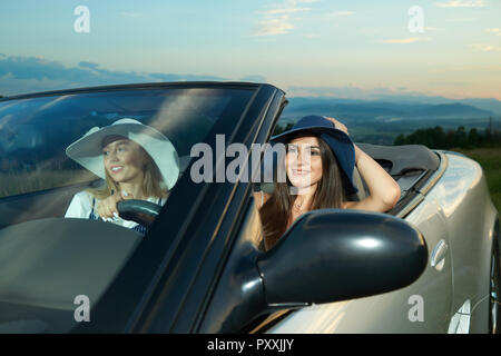 Piuttosto modelli in blu scuro e bianco orlo ampi cappelli seduti sui sedili anteriori di argento cabriolet. Adorabili ragazze auto pilotaggio, guardando lontano e sorridente. Concetto di automobile di lusso e di viaggio. Foto Stock