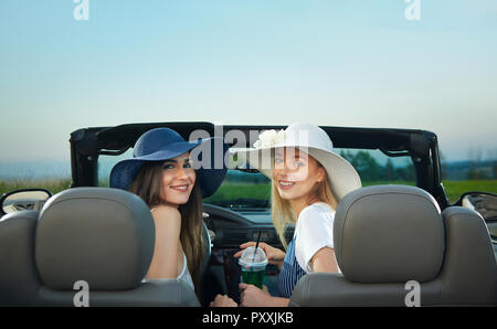 Vista dal retro di due giovani donne seduti sui sedili anteriori del cabriolet. Affascinante bruna e modelli bionda guardando indietro alla fotocamera e sorridente. Le ragazze che indossano in bianco e blu scuro orlo ampi cappelli. Foto Stock