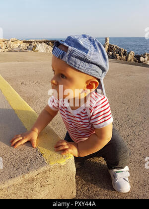 Carino Baby Boy 1 anno indossando un cappello indietro e vestiti a strisce, Close Up ritratto. Mare Mediterraneo in background Foto Stock