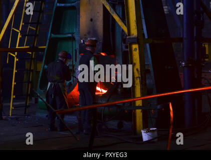 Fucinato metallico, forgiatura shop. Il martello idraulico forme red hot billetta. La produzione di componenti high-tech. Pezzi fucinati pesante impianto. Lavoratore mette la fattura Foto Stock