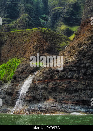 Bella cascata a cascata verso il basso il torreggiante pali (scogliere sul mare) visto da una barca in una giornata di sole, Costa di Na Pali, Kauai, Hawaii Foto Stock