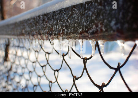 Riga diagonale di ghiaccioli appesi al recinto di maglia sotto lo strato di neve su un freddo gelido giorno in inverno Foto Stock