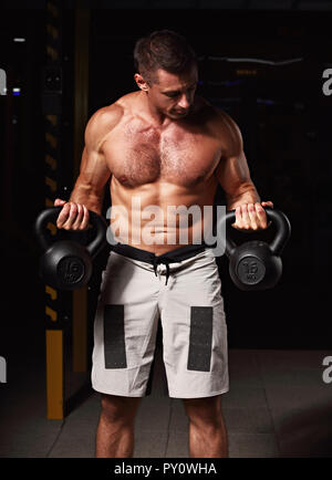 Forte uomo atletico facendo esercizi con kettlebells su ombra scura sullo sfondo. Closeup ritratto Foto Stock