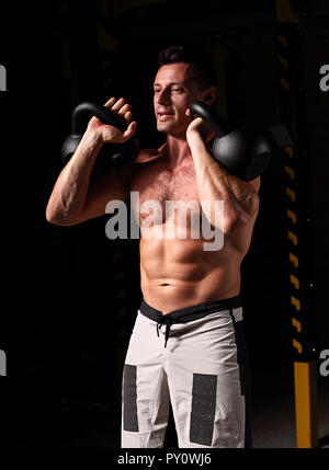 Bel uomo forte di fare gli esercizi con kettlebells su ombra scura sullo sfondo. Closeup ritratto di contrasto. Foto Stock
