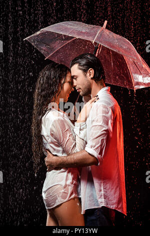 Vista laterale della coppia romantica in camice bianco con ombrellone in piedi sotto la pioggia su sfondo nero Foto Stock