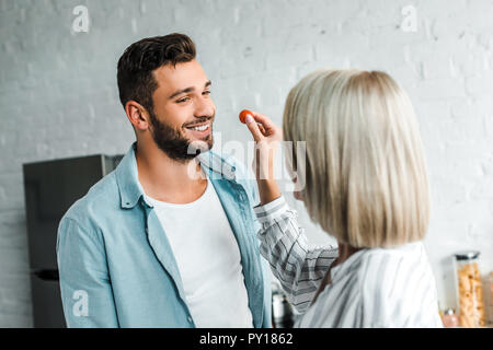 Ragazza sorridente alimentazione bello ragazzo con pomodoro ciliegino in cucina Foto Stock