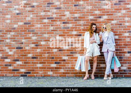 Bella giovani donne con le borse della spesa utilizza lo smartphone mentre in piedi di fronte a un muro di mattoni Foto Stock