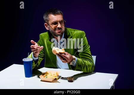 Uomo in giacca in velluto mangiare hamburger a tavola francese con grida e soda drink con sfondo blu Foto Stock