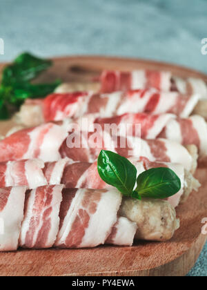 Freddo preparato per grigliare i suini salsicce avvolto in pancetta sulla piastra. Salumi crudi avvolto in pancetta servito fresco il verde del basilico foglie sul tagliere di legno. DOF poco profondo. Copia spazio per il testo. Foto Stock