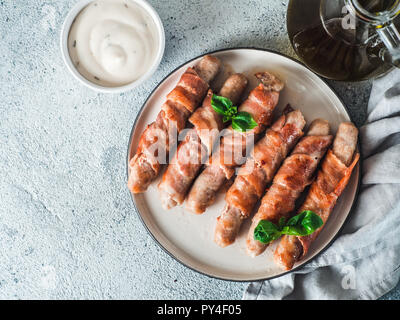 Pronti da consumare insaccati suini avvolto in pancetta sulla piastra. Fritti gustosi insaccati avvolto in pancetta servito fresco verde foglie di basilico con salsa di sfondo. Copia spazio per il testo. Vista superiore piatta o laici. Foto Stock