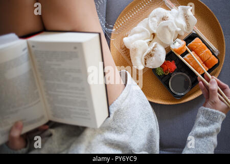 Giovane donna libro di lettura e mangiare sushi sul divano di casa. Vista aerea Foto Stock