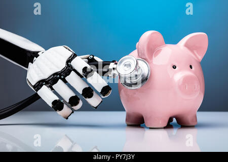 Close-up di una mano robotica esaminando Piggybank con uno stetoscopio Foto Stock