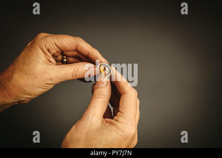 Close up di gioielli femminili maker mani esaminando l'anello in argento con pietra preziosa contro uno sfondo grigio. Gioielliere ispezione anello. Foto Stock