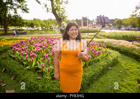 Donna asiatica tenendo selfie con telefono cellulare allega a selfie stick in posizione di parcheggio Foto Stock