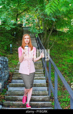 Una bella i Capelli rossi ragazza va giù per le scale e beve caffè da un bicchiere di carta. La prima colazione di stile di vita nel parco Foto Stock