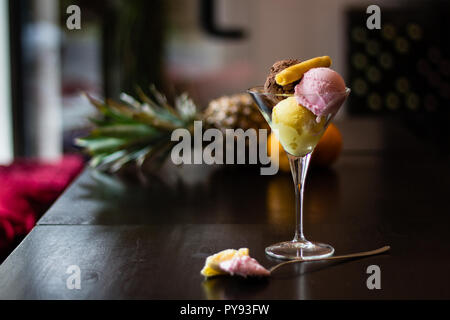 Palline di gelato, vaniglia, fragola, cioccolato in vetro su un tavolo di legno nella caffetteria decorata con cookie e ananas Foto Stock