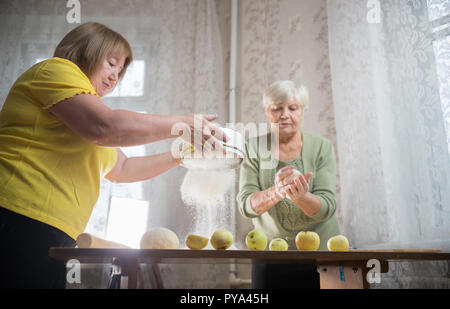 Due anziani donna rendendo poco apple pies a casa. Setacciare la farina sul tavolo Foto Stock