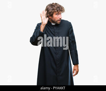 Bello ispanica sacerdote cattolico uomo sullo sfondo isolato sorridente con la mano sul orecchio in ascolto di un audizione di rumor o gossip. Concetto di sordità. Foto Stock