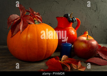 Due zucche con Bright Foglie di autunno apple, uve caraffa di rosso e blu sulla candela rustico in legno tabella contro un nero calcestruzzo ruvido grunge parete. Autunno Foto Stock