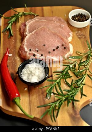 Raw non cotte fresche a fette di filetto di maiale piatto con rosmarino, pepe, sale, peperoncino rosso sul pannello di legno e sfondo nero. Vista laterale superiore, verticale Foto Stock