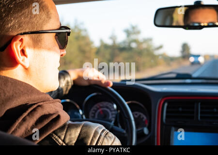 Uomo alla guida di una vettura in zona rurale, backseat vista. Driver maschio in un moderno veicolo su strada in un pomeriggio soleggiato Foto Stock