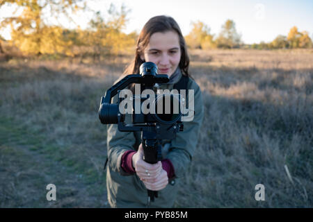 Videografo femmina tenendo una sospensione cardanica con fotocamera mirrorless. Donna con telecamera stabilizzata rig riprese in esterno in un pomeriggio soleggiato Foto Stock