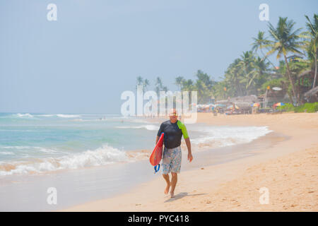 Hikkaduwa Lo Sri Lanka. Marzo 06, 2018. Un surfista passeggiate lungo la spiaggia con una tavola da surf. Foto Stock