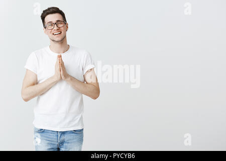 Prestare me un po' di soldi amico. Ritratto di affascinante sincero e carino giovane maschio brunet in bicchieri sorridendo tenendo le mani in preghiera sul petto, chiedendo il permesso o favore, in posa su uno sfondo grigio Foto Stock